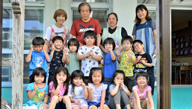 玉城善徳園長（後列左から２人目）と笑顔を見せる園児、職員＝１３日、沖縄市・室川保育園