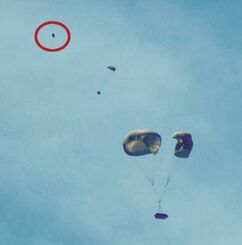 パラシュートで降下する物資と、パラシュートなしで落下するパネル状の物体（左上、丸で囲った部分）＝５日午前１１時２１分、うるま市の津堅島訓練場水域