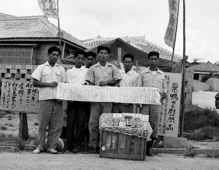 陳情小屋の前で、届いた慰問品と記念撮影する伊江島の住民。右端は阿波根昌鴻さん＝那覇市（わびあいの里提供）