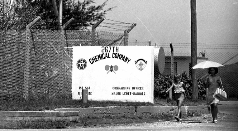 米軍の第２６７化学中隊と書かれたコンクリート製看板の前を横切る住民ら。生活の近くに毒ガスを貯蔵する施設があった＝１９６９年７月、美里村（現沖縄市）