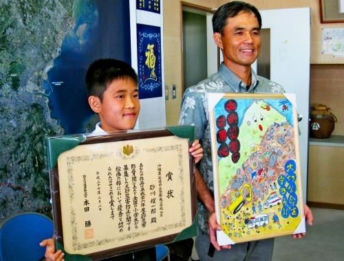 賞状と作品を手に、受賞を喜ぶ砂川耀一郎君（左）と父の徳久さん＝２５日、県庁